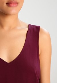 Burgundy ärmlös topp med djup V-ringning, mjuk textur och rena sömnadsdetaljer. Fokusera på tygdrapp och figurnära form.