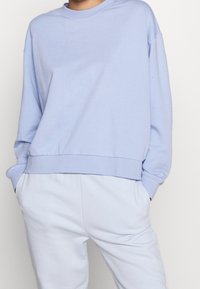 Felpa di cotone azzurro chiaro con collo tondo, spalle scese e orlo a costine. Abbinata a pantaloni della tuta azzurro chiaro.