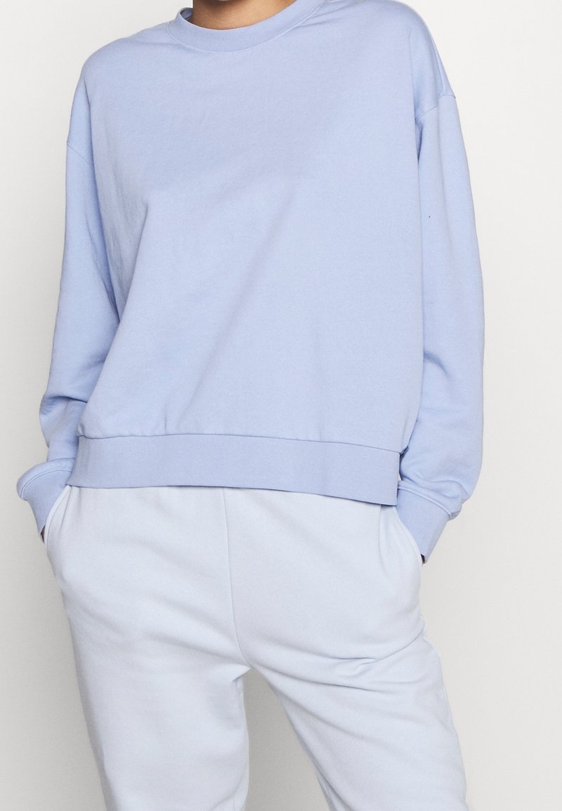 Felpa di cotone azzurro chiaro con collo tondo, spalle scese e orlo a costine. Abbinata a pantaloni della tuta azzurro chiaro.