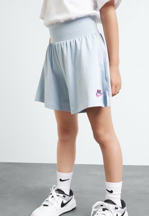 Barn iført lyseblå Nike-shorts, hvide Nike-sokker og hvide Nike-sneakers står på en grå gulv med en ensfarvet baggrund.
