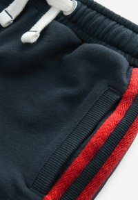 Marineblaue Sweatpants mit weißen Kordeln, ausgestattet mit roten Seitenstreifen und einem strukturierten Bund. Enthält ein seitliches Taschendetail.