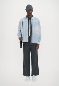 Lys blå denimjakke over en mørk skjorte, sammen med brede mørke bukser og hvite sneakers. En grå cap og en skulderveske fullfører looket.