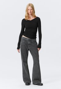Sort langærmet top med krydsning ved halsen, parret med højtaljede grå jeans med brede ben. Sorte flade sko fuldender outfittet.