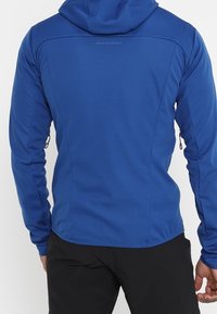 Giacca con cappuccio blu realizzata in tessuto liscio e elasticizzato, dotata di tasche con zip, linee di cucitura sagomate e una superficie leggermente texturizzata.