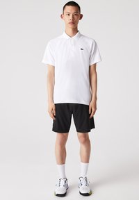 Vit pikétröja med korta ärmar och en grön logga, tillsammans med svarta shorts. Modellen har på sig vita strumpor och sportiga sneakers.