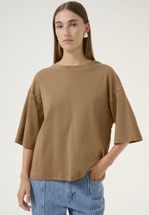 Brun bomulds t-shirt med korte ærmer og dekorative metalløjehuller på skuldrene, båret sammen med lyseblå jeans.
