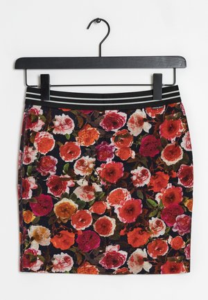 Jupe noire avec un motif floral vif en rouge, orange, rose et blanc, dotée d'une ceinture élastique rayée noire et blanche, suspendue à un cintre noir.