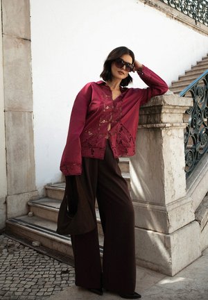 Blusa bordeaux con dettagli in pizzo, vestibilità oversize e maniche lunghe, abbinata a pantaloni larghi scuri, indossata di fronte a uno sfondo di scale in pietra.