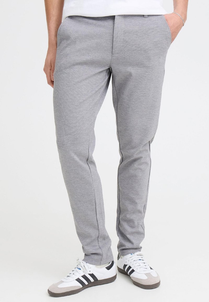 Άνδρας φορώντας ανοιχτογκρι slim-fit παντελόνια και λευκά αθλητικά παπούτσια με μαύρες ρίγες, στέκεται μπροστά σε λευκό φόντο.