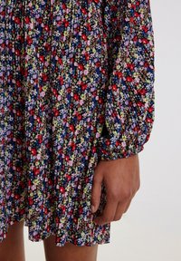 Blumenkleid aus dunklem Stoff mit Falten. Verziert mit roten, lila und grünen Blumen auf schwarzem Grund. Geraffte Manschetten und weiche Textur.