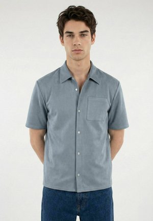 Ung mann iført en kortermet, lysegrå skjorte med knappelukking og brystlomme, og blå jeans, stående med hendene bak ryggen.