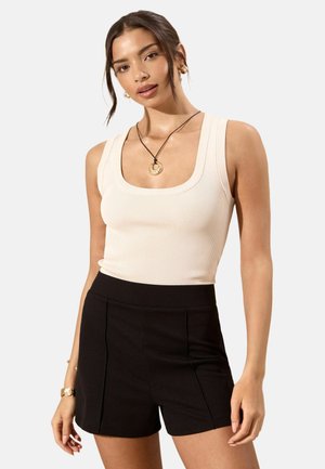 Frau mit braunen Haaren, die ein weißes, ärmelloses, geripptes Top, schwarze Shorts mit hoher Taille, goldene Creolen, eine Halskette und ein Armband trägt, steht vor einem weißen Hintergrund.