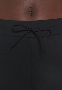 Gros plan sur une taille à peau foncée portant un pantalon noir avec un cordon de serrage noué au centre devant, sous le nombril.
