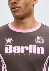 Κοντινό πλάνο ενός ατόμου που φοράει καφέ πουκάμισο με ροζ πατιλέτα και μεγάλο ροζ κείμενο που γράφει "Berlin" και γραφικά έμβλημα στο στήθος.