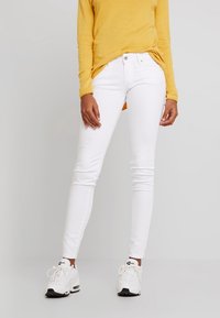 Los jeans ajustados blancos presentan una textura suave y un diseño entallado. La modelo los combina con una blusa amarilla de manga larga y zapatillas blancas.