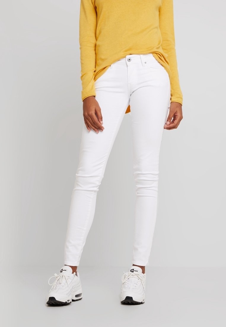 Los jeans ajustados blancos presentan una textura suave y un diseño entallado. La modelo los combina con una blusa amarilla de manga larga y zapatillas blancas.