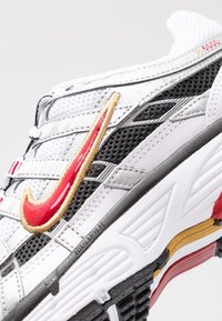 Bílé sportovní boty s černou síťovinou, červenými a zlatými akcenty, s výraznou Nike swoosh a texturovanou gumovou podrážkou. Kontrastní designové prvky.