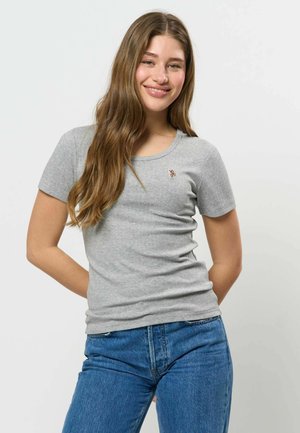 Jeune femme aux longs cheveux bruns souriante, portant un t-shirt gris clair à manches courtes et un jean bleu, debout devant un fond clair uni.