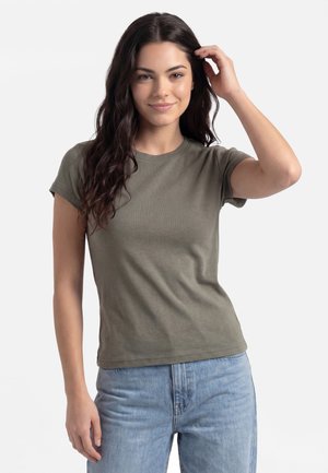 Ung kvinde med langt mørkt hår iført en olivengrøn t-shirt og lyseblå jeans, smilende og rører ved sit hår med den ene hånd.