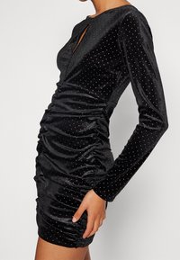 Robe en velours noir à manches longues, côtés froncés et ornée de clous argentés pour la texture. Détail d'encolure en trou de serrure.