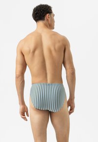 Ein Mann mit lockigem Haar steht mit dem Rücken zur Kamera, trägt hellblaue und braune vertikale gestreifte Unterwäsche und steht vor einem schlichten weißen Hintergrund.