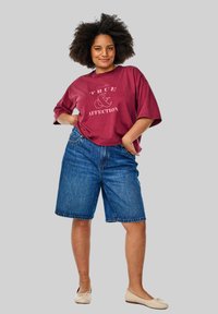 Burgundijas oversize T-krekls ar baltu tekstu; zili denima šorti ar brīvu fit. Beigādas kurpes pabeidz apģērbu. Pelēks fons.