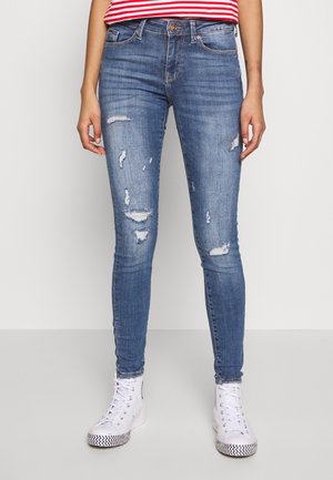 Bas du corps portant un jean skinny bleu déchiré, des baskets blanches montantes et une chemise rayée rouge et blanche.