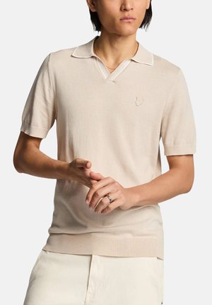 Giovane che indossa una polo beige a maniche corte con un piccolo logo ricamato sul petto, con le mani unite davanti.