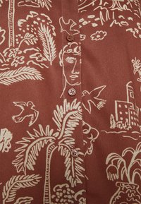 Tissu marron avec imprimé crème représentant un visage humain tropical et abstrait, avec des palmiers, des oiseaux, des bâtiments et une fermeture à boutons au centre.
