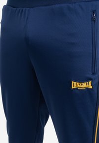 Pantalons de jogging de sport bleu marine en tissu lisse, dotés de poches latérales zippées et d'un logo Lonsdale jaune sur la cuisse gauche.