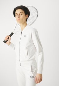 Balts sporta treniņtērps ar rāvējslēdzēja jakas un atbilstošām biksēm. Gluda tekstūra; uz jakas un biksēm ir logotips. Tur rokās tenisa raketi.