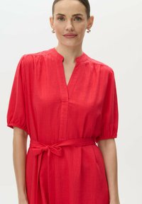 Czerwona sukienka z krótkim rękawem, posiadająca kołnierz mandaryński i wiązanie w pasie. Wykonana z teksturowanej tkaniny, ma luźny krój i marszczone detale na rękawach.