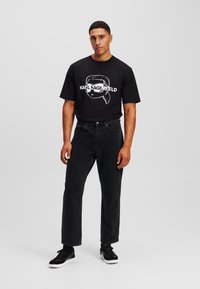 Camiseta de algodón negra con una impresión blanca de un personaje con gafas de sol y el nombre "KARL LAGERFELD". Combinada con jeans oscuros y zapatillas negras.