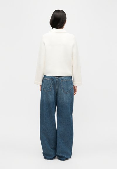 Γυναίκα με ίσια σκουρόχρωμα μαλλιά που φοράει λευκό μακρυμάνικο μπουφάν και φαρδιά μπλε τζιν παντελόνια, στέκεται γυρισμένη προς τα πίσω σε απλό φόντο.