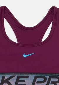 Fialová športová podprsenka Nike Pro s modrým logom Nike swoosh a širokým elastickým pásom s textom "NIKE PRO" v čiernej farbe.