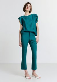 Donna in piedi che indossa una blusa azzurro mare a maniche corte con maniche a ruffles, pantaloni slim coordinati, sandali con tacco aperti bianchi e orecchini a cerchio in argento.