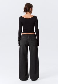 Femme avec les cheveux attachés en chignon, portant un haut noir à manches longues et un jean noir à jambes larges, debout de dos sur un fond clair uni.