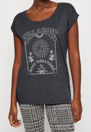 Person iført mørkegrå t-shirt med "Billabong Good Vibrations" og grafik med sol, yin-yang, blomster, måne og palme, kombineret med mønstrede bukser.