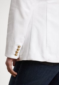 Λευκό σακάκι με υφασμάτινη υφή, μονή σχισμή στο πίσω μέρος και τέσσερα χρυσά κουμπιά στη μανσέτα. Συνδυάζεται με σκούρο τζιν παντελόνι.