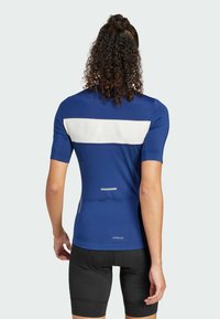 Maillot de cyclisme bleu à manches courtes, avec une bande horizontale blanche dans le dos et des accents réfléchissants. Un short de cyclisme noir est porté.