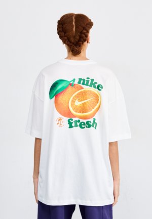 Camiseta blanca de gran tamaño con un gráfico de una naranja con una hoja verde, texto "nike" y "fresh" en verde, y un pequeño detalle de logo debajo.