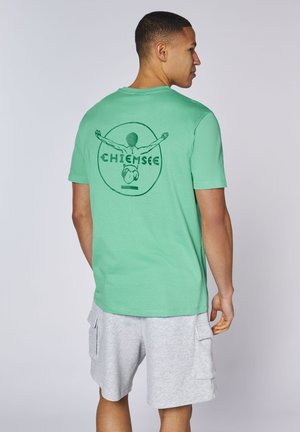Mann trägt grünes T-Shirt mit Schwimmer-Grafik und dem Schriftzug „Chiemsee“ auf dem Rücken, kombiniert mit hellgrauen Cargoshorts, steht vor schlichtem Hintergrund.