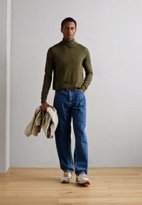 Olivově zelený rolák, modré džíny a béžová vnitřní bunda, doplněné bílými a oranžovými teniskami, stojící na dřevěné podlaze.