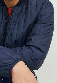 Quiltad marinblå bomberjacka med ribbad krage och manschetter. Har en frontdragkedja och textil med struktur för värme och komfort.