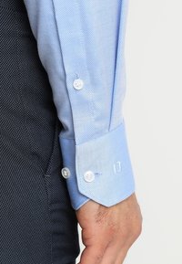 Camicia a quadri blu chiaro con polsini abbottonati, bottoni bianchi e una texture liscia; abbinata a un tessuto scuro e testurizzato sotto.