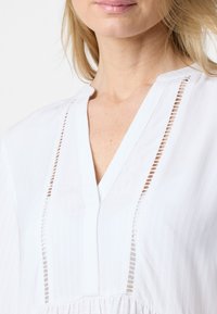Weiße Bluse mit vertikalen Streifen, mit V-Ausschnitt, Ausschnitten und subtiler Textur. Weiche Stoffzusammensetzung und gerafftes Saumdetail.