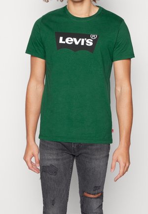 Osoba ubrana w zielony t-shirt Levi's i czarne, podarte jeansy, stojąca na tle jasnego, jednolitego tła.