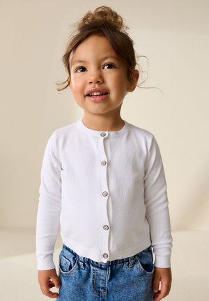 Lächelndes junges Kind mit braunen Haaren in einem unordentlichen Dutt, trägt eine weiße, geknöpfte Strickjacke und blaue Jeans, steht vor einem schlichten Hintergrund.