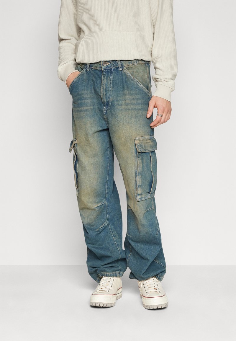 Stora blå denim cargo-byxor med sidofickor, blekt utseende, avsmalnande anklar och subtila slitmönster, tillsammans med vita sneakers.