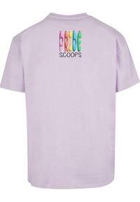 Laventelinvärinen lyhythihainen T-paita, jossa on monivärinen "PRIDE" -grafiikka ja musta "SCOOPS" -teksti yläselässä. Pehmeää puuvillamateriaalia.